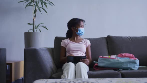 African american girl wearing face mask using digital tablet while sitting on the couch at hospital