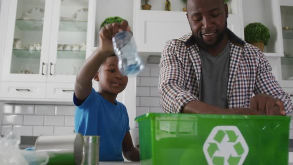 Happy african american father and son standing in kitchen putting plastic rubbish in recycling box