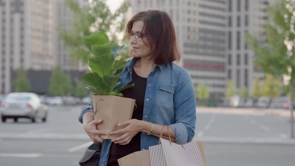 Middle Aged Woman with Paper Shopping Bags with Buying Plant Looking at Houseplant