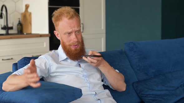 Charismatic Redhead Guy Record Voice Message and Smiling Sits on the Sofa at Home