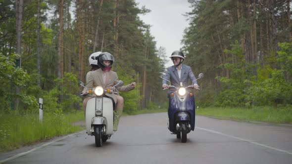 Confident Scooter Drivers Bumping Wrists As Riding Along Suburban Road on Cloudy Summer Day. Wide