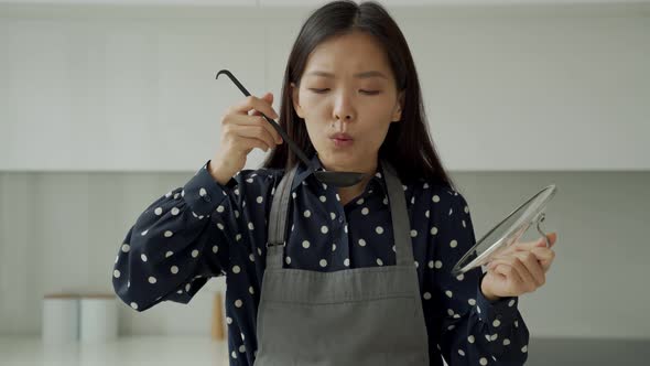 Asian Woman in Gray Apron Tries Hot Soup with a Spoon in the Kitchen and Smiles at the Camera