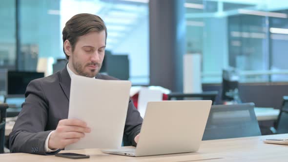 Businessman with Laptop Reading Documents in Office