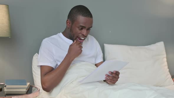 African Man Celebrating Success on Documents in Bed