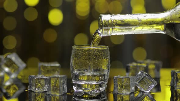 Barman Pour Frozen Vodka From Bottle Into Shot Glass. Ice Cubes Against Shiny Gold Party Background