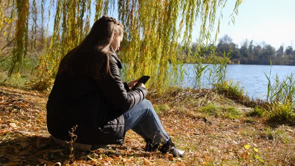 Young Woman with a Smartphone in Hands Sits on Yellow Foliage By River in Autumn