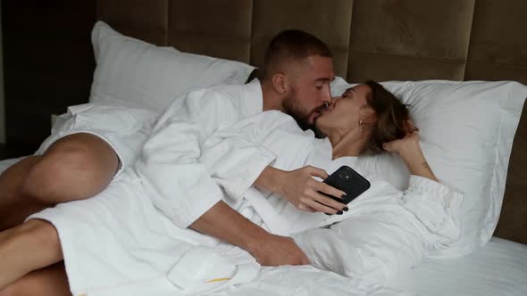 Morning in Hotel Room Man and Woman are Embracing in Bed Lady is Using Smartphone