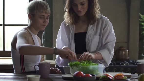 Beautiful Homosexual Women Talking and Tearing Green Salad at Table in Home Kitchen