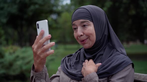 Positive Senior Woman in Hijab Talking Using Video Chat on Smartphone Standing in Park Outdoors