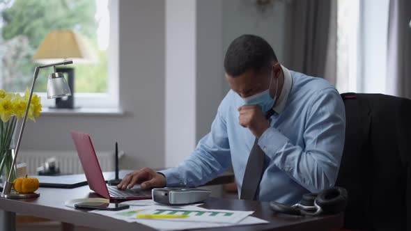 Young Focused African American Man in Coronavirus Face Mask Coughing Messaging Online and Using