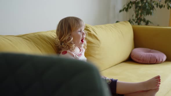 Little Happy Cheerful Girl Child in Pink Dress Sits on Floor in Living Room on Yellow Sofa and