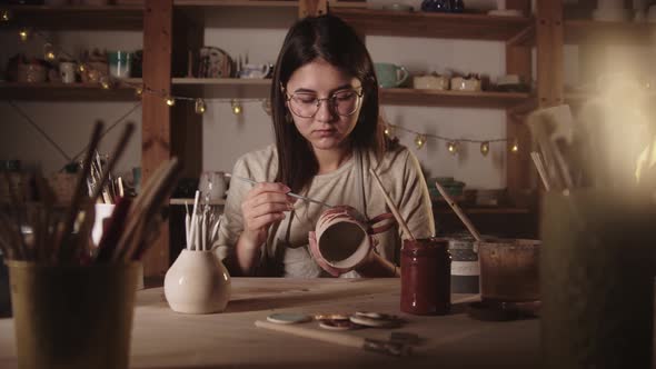 Young Woman Potter in Glasses Drawing a Design on the Ceramic Mug with a Brush
