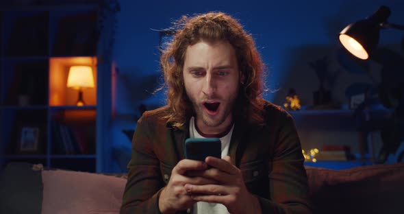 Cheerful Hipster Guy Making Surprised Face While Looking at Phone Screen, Handsome Long Haired Man