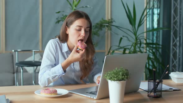 Young Businesswoman Eating a Glazed Donut While Working on a Laptop at the Office Table