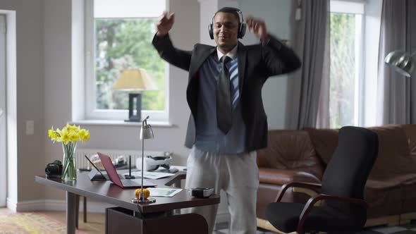 Joyful African American Young Man in Headphones Dancing in Home Office Indoors