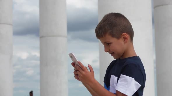 Tanned Boy is Watching at Phone in His Hands on the Background of White Columns