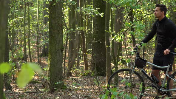 A Cyclist Stands with a Bicycle in a Forest and Looks Around, Lost