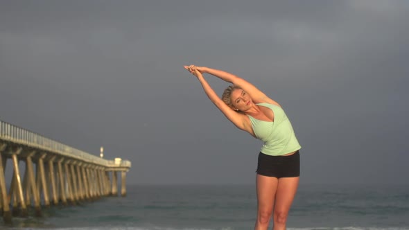 A young woman does yoga on the beach next to a pier.