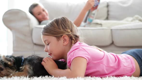 Little girl playing with puppy with her mother reading on the sofa