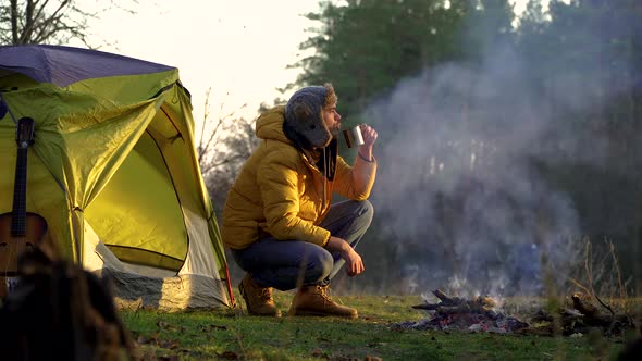 A Man Drinks Tea Near a Campfire and a Tent in the Woods