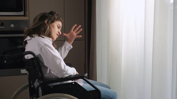 Pensive Disabled Woman Sits in Wheelchair By Window at Home