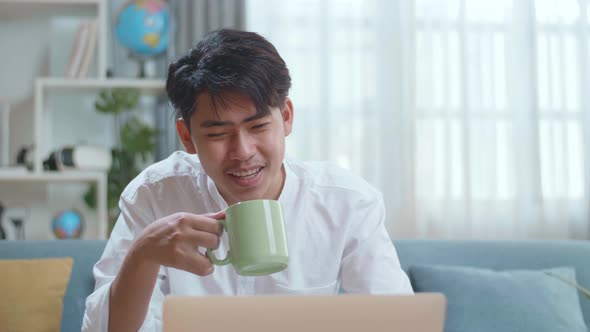 Happy Asian Man Drinking Coffee While Using Laptop Computer In The Living Room