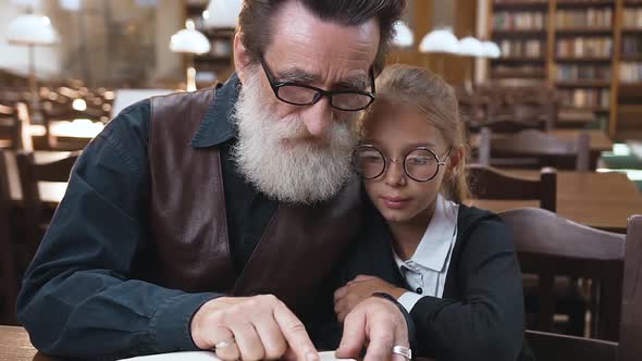 Grandfather with Well-Groomed Beard which Reading Book His Pretty Teen Granddaughter in the Library