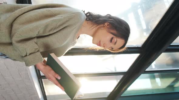 Beautiful Woman Sitting Near Window and Reading a Book on Background of Sunset