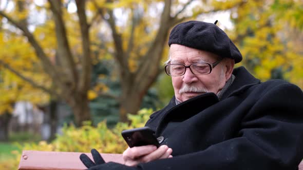 An Old Grandfather is Talking on a Smartphone He is Sitting on a Bench in the Park