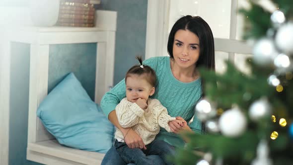 Mother Hugging Her Daughter Sitting on the Windowsill Near the Christmas Tree