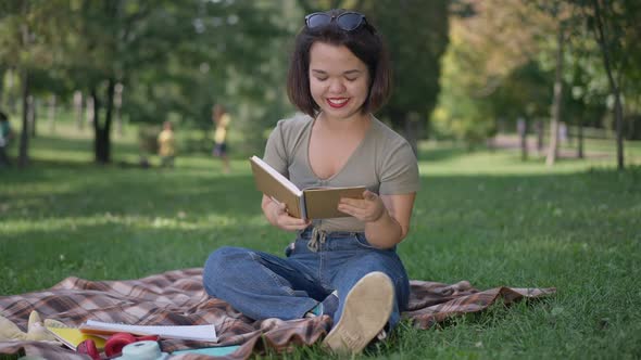 Wide Shot Portrait of Happy Little Woman with Dwarfism Reading Book Sitting on Blanket in Summer