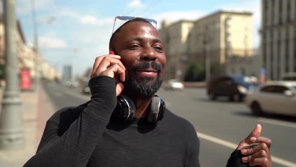 Black Man Is Walking in City at Sunny Day and Talking By Cell Phone, Strolling Along Road with Cars