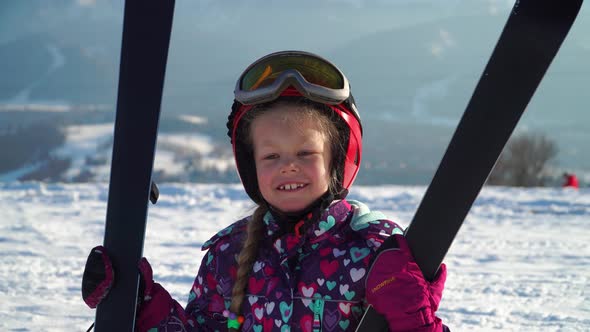 Little Girl Wearing Red Helmet and Outwear Holding Mountain Skis in Sunlight
