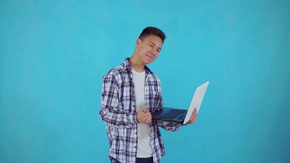 Joyful Young Asian Man with Laptop in Hand Learns About Winning on Blue Background