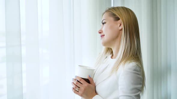 Happy Young Married Woman Drinks Tea and Day Dreams in Front of Window