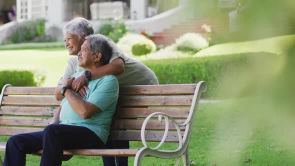 Video of happy biracial senior couple embracing and sitting on bench in garden