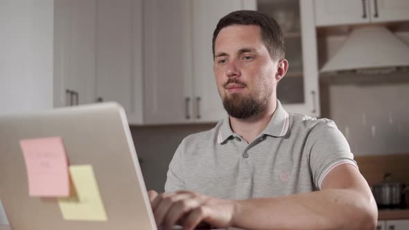 Adult Cheerful Man Is Working with Notebook in a Home, Sitting on a Kitchen