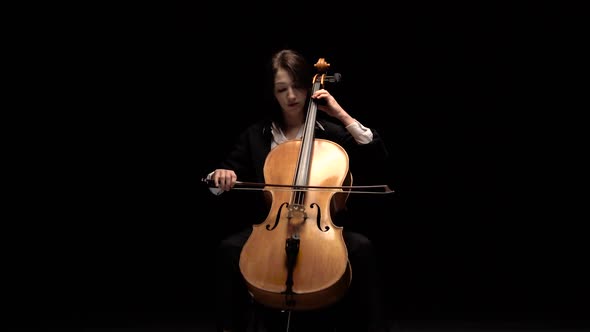 Girl Plays a Cello a Sitting on a Chair in a Dark Room. Black Background