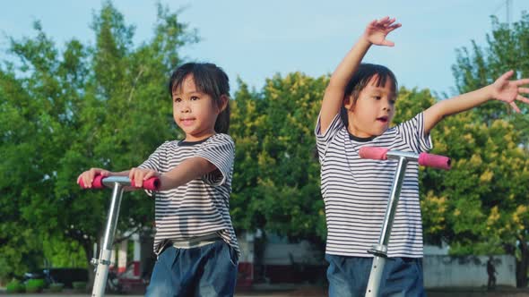 Two cute little girls riding scooter on road in park outdoors on summer day.