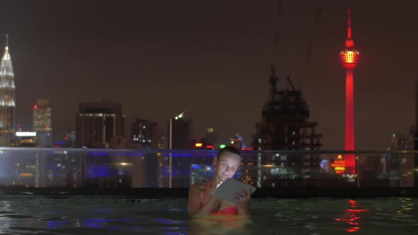 View of Woman in Swimming Pool on the Skyscraper Roof Using Tablet Against Night City Landscape