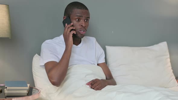 Angry African Man Talking on Smartphone in Bed