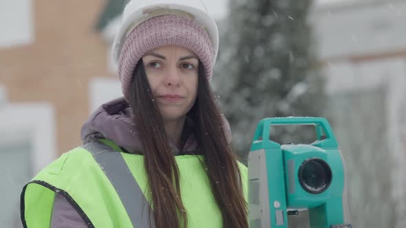 Closeup Portrait of Confident Female Surveyor Standing at Theodolite Gesturing on Winter Day