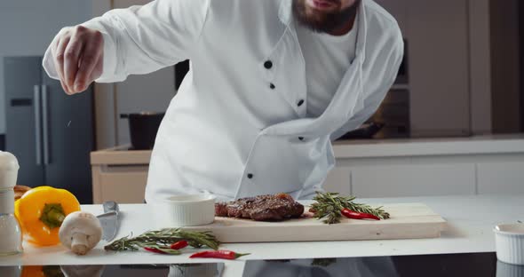 Chef in White Uniform Serving Grilled Beef Meat with Rosemary Twigs on Wooden Board