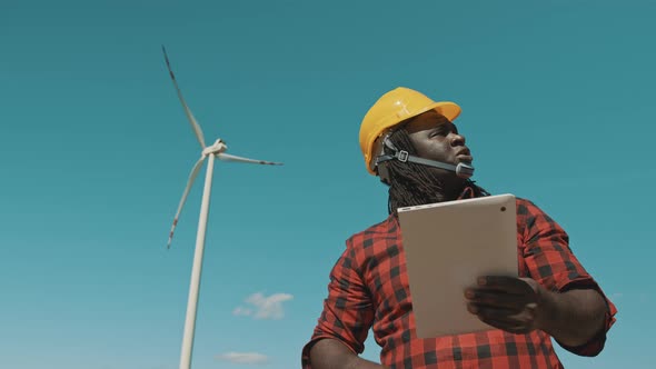 African Man in Hard Hat Standing with Modern Digital Tablet Against Windmill Power Station Making