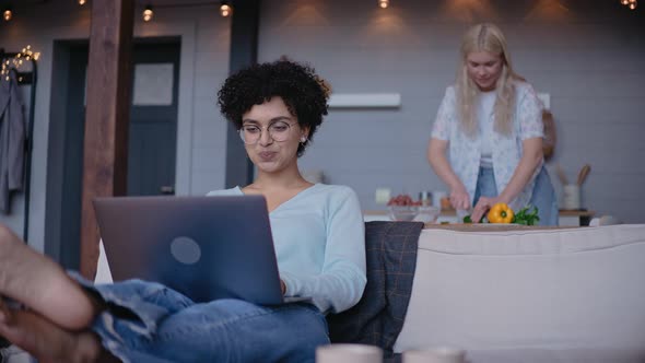 Young Girl Looks at the Laptop and Talks with Her Girlfriend Cooking Behind Her