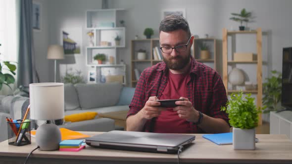 Happy Man Playing Game on Smartphone Sitting By Table at Home