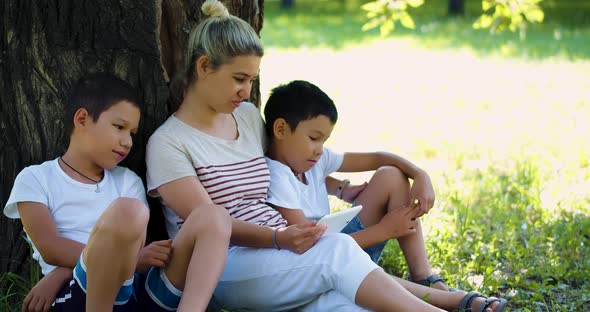 Footage of an Asian Woman with Two Children Watching Online Video While Sitting Under a Tree in the