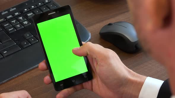 An office worker scrolls at a smartphone with a green screen at a desk with a computer - closeup