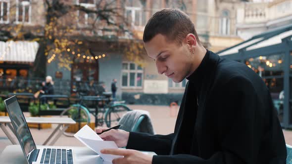 Young Attractive Businessman Sits in Cafe Reading Paper Documents.