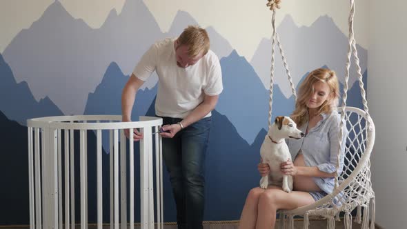 a Man Collects a Crib for a Child, His Wife Plays with a Dog Jack Russell Sitting on a Swing